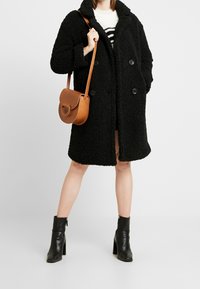 Černý, strukturovaný, dvouřadový kabát s velkými knoflíky; doplněný hnědou kabelkou přes rameno a černými kotníkovými botami. Pruhovaný svetr pod ním.