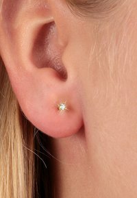 Goldfarbene Steckerohrringe mit einem kleinen, klaren Edelstein, vor einem Nahaufnahme von einem leicht rosafarbenen Hautton und einem Ohr. Funkelnde Details sind sichtbar.