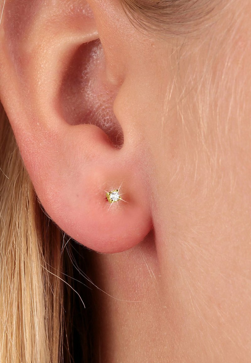 Goldfarbene Steckerohrringe mit einem kleinen, klaren Edelstein, vor einem Nahaufnahme von einem leicht rosafarbenen Hautton und einem Ohr. Funkelnde Details sind sichtbar.