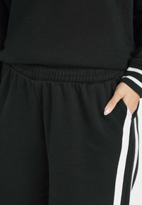 Pantalons de jogging noirs avec une taille élastique, présentant des rançons blanches sur les côtés et des poches latérales, fabriqués en tissu doux et texturé.
