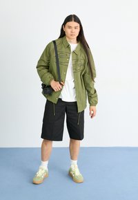 Olive green zip-up jacket with black outlines, white t-shirt, black shorts, and green sneakers with light yellow laces. Compact black bag.