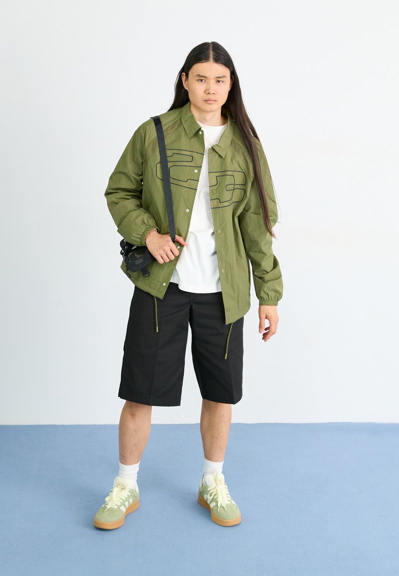 Giacca a zip verde oliva con bordi neri, t-shirt bianca, pantaloni shorts neri e sneakers verdi con lacci giallo chiaro. Borsa nera compatta.