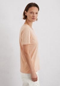 Light peach-colored t-shirt made of soft, textured fabric. Features short sleeves and a round neckline. Worn with white pants.