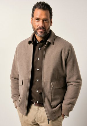 Hombre de mediana edad con barba, vestido con una chaqueta beige de cremallera sobre una camisa marrón oscuro y pantalones beige, de pie con las manos en los bolsillos.