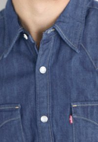 Denim shirt in dark blue with a pointed collar, two chest pockets, white buttons, and yellow stitching accents.