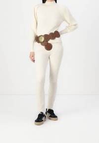 Brązowy skórzany pasek z okrągłymi akcentami i centralnym ozdobnym elementem, noszony na jasnym kremowym swetrze w prążki i jasnych spodniach. Czarne trampki.