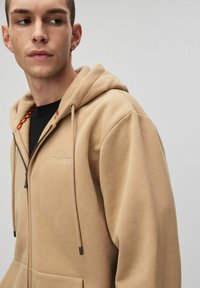 Beżowa bluza z kapturem zapinana na zamek, wykonana z mieszanki bawełny, z teksturowanym wykończeniem, regulowanymi sznurkami oraz haftowanym logo na klatce piersiowej.