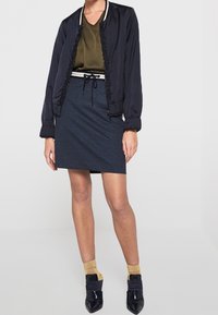 Laivastonsininen bomber-takki, jossa röyhelöreunus, oliivinvihreä yläosa, tummat farkkuhame ja mustat korkokengät. Kultakoristetut sukat lisäävät tekstuuria ja kontrastia.