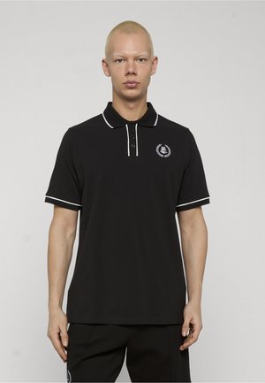 Młoda osoba o krótkich blond włosach, ubrana w czarną koszulkę polo z białym obszyciem i małym emblematem na lewej piersi, stojąca na jednolitym tle.