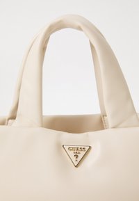 Sac en cuir doux couleur crème avec des poignées rembourrées et une petite plaque triangulaire dorée avec le logo Guess USA sur le devant.