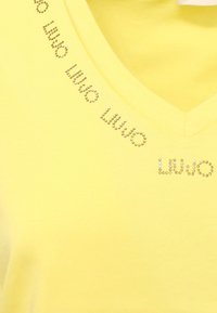 T-shirt giallo a V in morbido tessuto, con un motivo decorativo di borchie argentate disposte in un design di logo attorno al colletto.