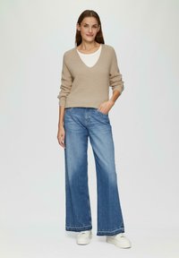 Beige Strickpullover mit V-Ausschnitt, kombiniert mit weit geschnittenen blauen Jeans. Weiße Sneakers, lässiger Look. Leichte Textur, entspannter Schnitt und bequeme Optik.