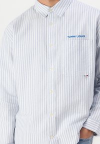 Jasnoniebieska i biała koszula z guzikami w paski, z kieszonką, z haftem "TOMMY JEANS" w kolorze niebieskim oraz małą metką z logo na rękawie.