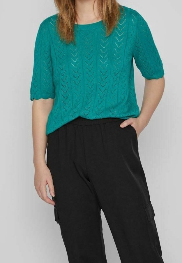 Femme portant un haut en maille à manches courtes couleur sarcelle avec un motif chevron et un pantalon noir taille haute, debout avec une main dans la poche.