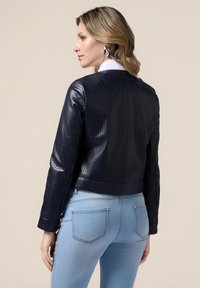 Giubbotto in pelle traforata color navy con silhouette aderente e polsini a coste, indossato su jeans attillati azzurri. Texture liscia e leggero luccichio.