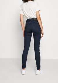 Jean skinny bleu foncé taille haute avec une texture denim lisse, dotés de poches arrière, associés à un t-shirt blanc et des baskets blanches.