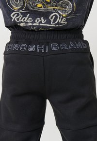 Detail zblízka na pás s čiernymi nohavicami s textom "KOROSHI BRAND" a čiernu košeľu so žltým motocyklem a sloganom "Ride or Die".