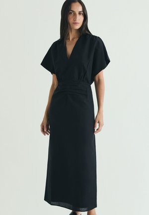 Mujer de pie con un vestido largo negro con mangas cortas y anchas y un escote en V pronunciado, sobre un fondo claro y liso.