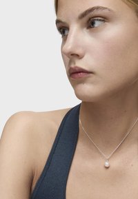 Srebrny naszyjnik z małym zawieszeniem z perłą, z delikatnym łańcuszkiem. Modelka nosi ciemny, strukturalny top bez rękawów.