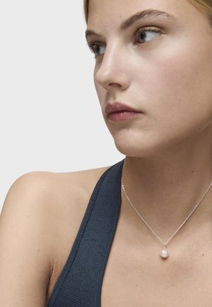 Srebrny naszyjnik z małym zawieszeniem z perłą, z delikatnym łańcuszkiem. Modelka nosi ciemny, strukturalny top bez rękawów.