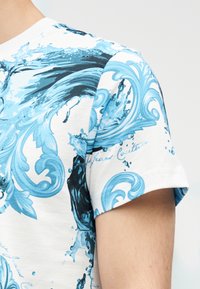 Κοντομάνικο λευκό πουκάμισο με μπλε και μαύρα κυματιστά και σπειροειδή σχέδια στον ώμο και το πάνω μέρος του βραχίονα.