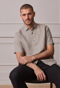 Hellbeiges Kurzarmhemd mit Kragen, Knopfleiste vorne und strukturierter Stoff. Getragen mit dunkler Hose, sitzend auf einem Stuhl.