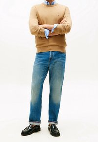 Homme portant un pull camel par-dessus une chemise bleue à rayures, un jean bleu avec les ourlets retroussés, et des mocassins en cuir noir, debout les bras croisés.