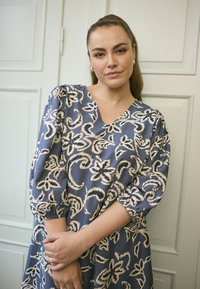 Modrá kvetinová šaty s výstrihom do V, vypchatými rukávmi a elastickými manžetami. Tkanina má hladkú textúru s kontrastnými béžovými vzormi.