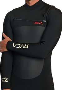 Mann in einem schwarzen RVCA-Neoprenanzug mit einer Reißverschlusstasche auf der Brust und Logos auf Brust und Ärmeln, die Arme über dem Oberkörper verschränkt.