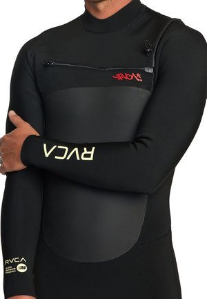 Uomo che indossa una muta nera RVCA con una tasca con zipper sul petto e loghi sul petto e sulle maniche, braccia incrociate sul torso.