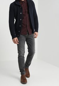 Mörkblå stickad cardigan med sjalkrage, knäppning, vinröd skjorta under, grå jeans och bruna läderstövlar.