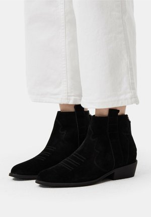Stivaletti neri in suede con punta affusolata, cuciture decorative e tacco a blocco basso, indossati con pantaloni bianchi a vita corta.