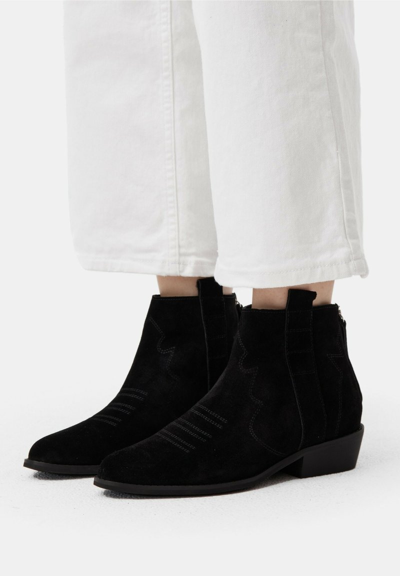 Botine negre din nubuc, cu vârful ascuțit, cusături decorative și toc mic, purtate cu pantaloni albi scurți.