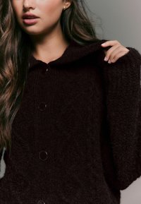 Dunkelbrauner Strickpullover mit Kapuze, der eine strukturierte Oberfläche, eine Knopfleiste mit großen schwarzen Knöpfen und überschnittene Schulternähte aufweist.