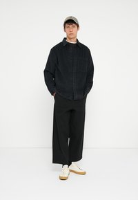 Black checkered shirt, loose fit, single chest pocket; paired with wide black pants and white sneakers with beige soles. Neutral cap worn.