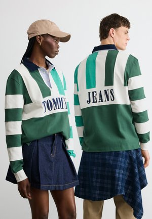 Két modell zöld-fehér csíkos rögbiingben, amelyeken a "TOMMY" és a "JEANS" felirat látható, az egyik farmer szoknyával, a másik kockás anyaggal párosítva.