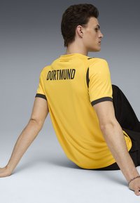 Geltona sportinė marškinė su juodais akcentais, ant nugaros užrašytas "DORTMUND", trumpomis rankovėmis ir lygiu paviršiumi. Matomos juodos kelnės.