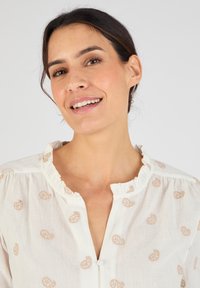 Blusa bianca con scollo arricciato, caratterizzata da motivi decorativi beige in tutto il capo. Tessuto leggero con una vestibilità morbida e chiusura a bottoni.