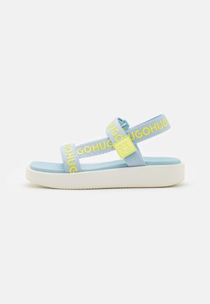 Sandalia de plataforma azul claro con suela crema, correas ajustables con texto amarillo "HUGO" y hebilla amarilla con marca en el lateral.