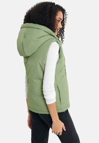 Grüne Pufferweste mit Kapuze, aus glattem Material, mit seitlichen Taschen und tailliertem Schnitt. Getragen über einem weißen Langarmshirt.