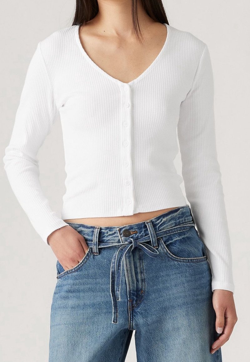 Femme portant un crop top blanc à manches longues côtelé avec boutons et un jean en denim bleu taille haute avec ceinture nouée, une main dans la poche.