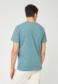 Maglietta a maniche corte color turchese con collo a giro, realizzata in un tessuto misto cotone; indossata in modo casual con pantaloni chiari.