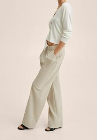 Person trägt beige weite Hosen, ein weißes, kurz geschnittenes Langarmshirt, schwarze Sandalen mit offenen Zehen, hat die Hände in den Taschen und steht vor einem einfarbigen Hintergrund.