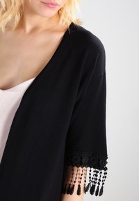 Musta huivi, jossa syvä V-kaula-aukko, lyhyet hihat, virkattua pitsiä ja helminyöritykset hihojen reunoissa. Pehmeä kangas.