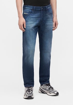 Homme portant un jean bleu délavé et des chaussures de sport argentées et noires, debout avec les bras détendus le long du corps.