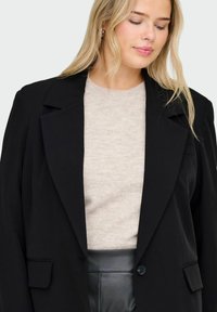 Melna žakete ar struktūru dizainu, viena pogas aizdare un divas priekšējās kabatas, uzvilkta virs gaiši bēša džempera. Gluda auduma tekstūra.
