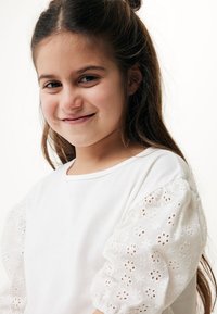 Camiseta blanca de manga corta abullonada hecha de tela de eyelet estampada. La camisa tiene un escote redondeado y un corte holgado.