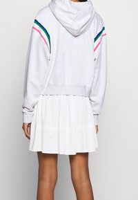 Person wearing a gray hoodie with blue, green, and pink stripes on sleeves and a white pleated skirt, standing with back facing camera.