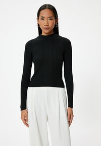 Schwarzes, geripptes Langarmshirt mit hohem Kragen und figurbetontem Design, kombiniert mit weit geschnittenen weißen Hosen. Minimale Akzente, glatte Textur.