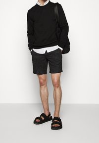 Suéter negro, camisa blanca con cuello, pantalones cortos a rayas negros y sandalias negras. El material parece casual con un ajuste y textura informales.